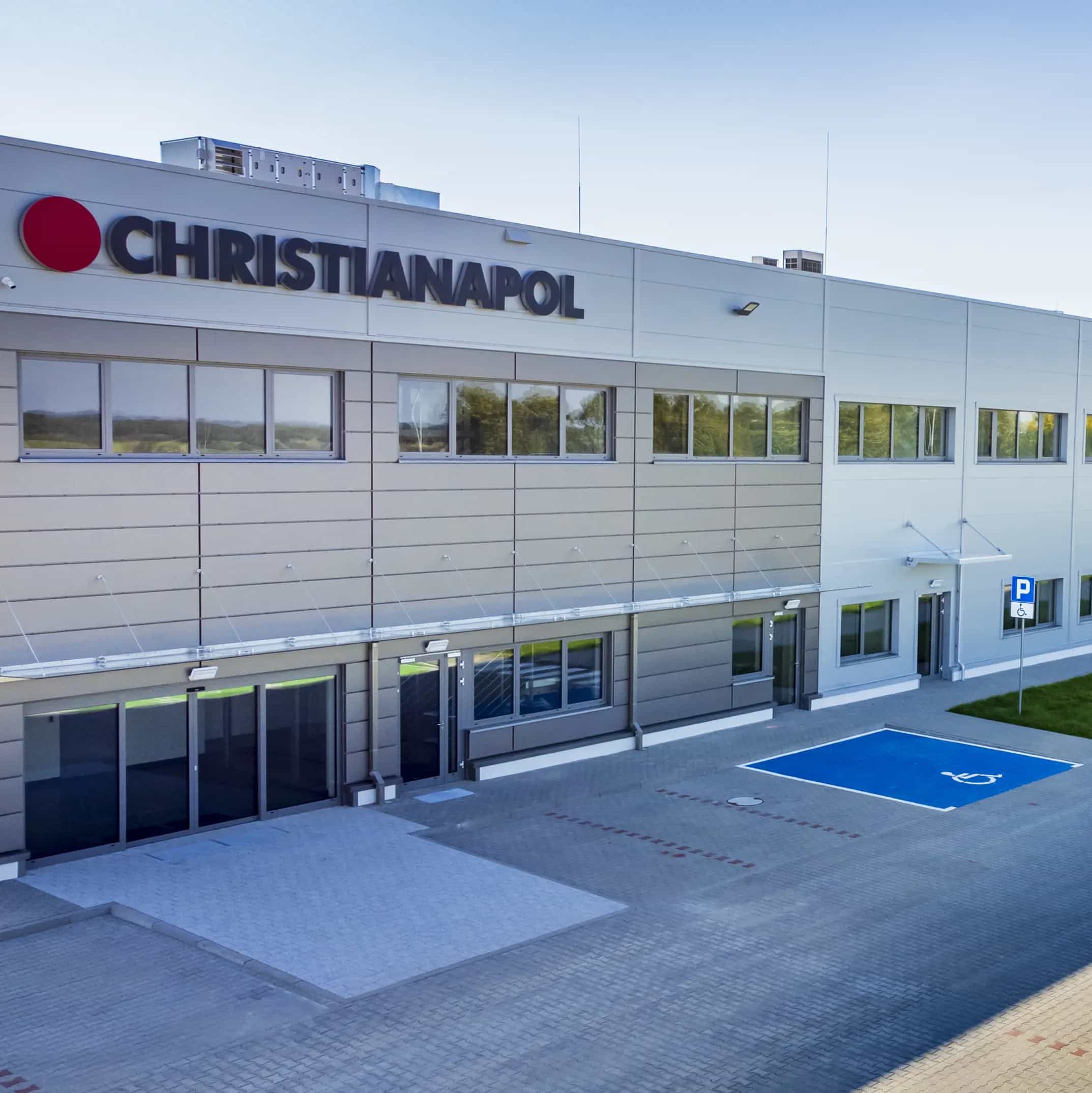Budynek przemysłowy z logo Christianapol, szeroki chodnik i oznaczone miejsca parkingowe na pierwszym planie.